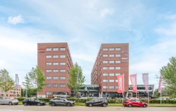 Twee moderne bakstenen kantoorgebouwen op Rijnzathe 12, De Meern, met voorzijde geparkeerde auto's en vlaggen. Ideaal voor kantoorruimte huren met goede zichtlocatie en toegang.