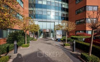 Toegang tot kantoorgebouw aan de Olof Palmestraat 10 in Delft, met moderne glazen entree omgeven door bakstenen gevel en groenvoorzieningen. Ideale locatie voor kantoorruimte huren.