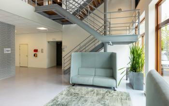 Ruime, lichte entree van een kantoorpand op Delftechpark 9, Delft. Grote ramen, grijze trap en moderne zitbank. Ideaal voor bedrijven die kantoorruimte willen huren met een professionele uitstraling.