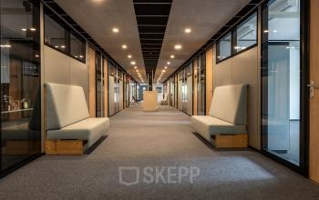 Lichte gang in modern kantoorpand op Delftechpark 9, Delft, met glazen kantoorruimten aan beide zijden en grijze vloertapijten. Benches langs de muren bieden zitmogelijkheden. Ideaal voor kantoorruimte huren.