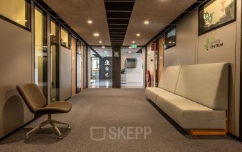 Kantoorruimte huren op Delftechpark 9 in Delft, met moderne grijze stoelen en banken in een ruime gang. Verlichte plafonds geven een heldere uitstraling aan de kantoorgang.