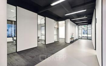 Moderne, strak vormgegeven kantoorruimte huren in Den Bosch met glazen wanden, zwart plafond en verlichting. Open gang die toegang biedt tot verschillende kantoren. Geen personen aanwezig.