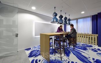 Moderne kantoorruimte in Den Bosch met een grote houten tafel en kleurrijk blauw vloerkleed. Twee mensen zijn in gesprek aan de tafel. Ideaal voor wie kantoorruimte wil huren.
