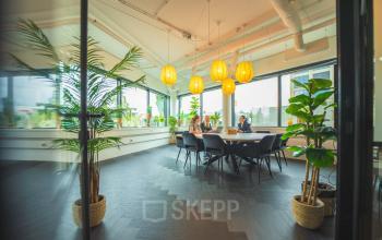 Lichte kantoorruimte met grote ramen, zes hanglampen en tafels met stoelen, gelegen op Hambakenwetering 1, Den Bosch. Twee personen zijn bezig met een zakelijke bespreking.