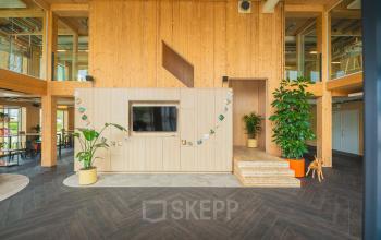 Ruimte in kantoorpand op Hambakenwetering 1-kr, Den Bosch. Houten wanden, planten en een ingebouwde tv geven een moderne uitstraling. Ideaal voor kantoorruimte huren. Mensen zitten ontspannen in een hoek.