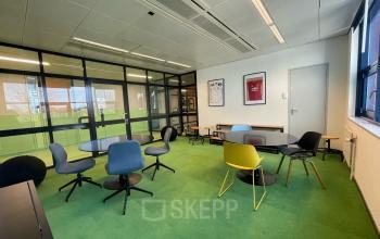 Een kleurrijk kantoorpand met glazen wanden en verschillende stoelen op groene vloerbedekking, gelegen op Bruistensingel 500 in Den Bosch. Ideaal om kantoorruimte te huren voor creatieve en zakelijke samenwerkingen.