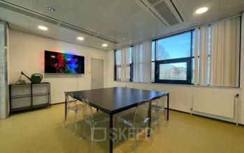 Kantoorruimte huren in Den Bosch, bij Bruistensingel 500. Heldere vergaderruimte met grote ramen, een lange tafel en stoelen, ideaal voor zakelijke bijeenkomsten in dit kantoorpand.
