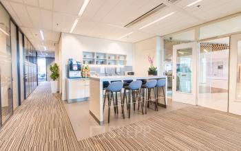 Moderne kantoorruimte te huur op Koningin Julianaplein 10, Den Haag Centrum. De heldere pantry heeft een koffiezetapparaat, barkrukken aan een hoge tafel en enkele decoratieve planten.