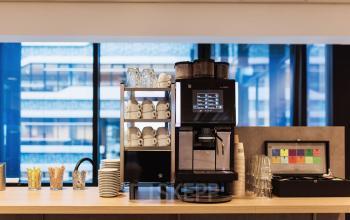 Moderne koffiehoek in een kantoorpand aan Anna van Buerenplein 41, Den Haag Centrum, met een koffiemachine, bekers en glazen tegen een glazen achtergrond.