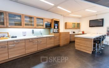 Moderne kantoorpantry in Den Haag Centrum met houten kasten, ingebouwde apparaten en een bar met zitplaatsen. Geschikt voor huur als praktische koffie- en lunchruimte.