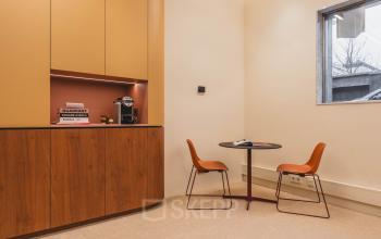 Kantoorruimte huren bij Zeestraat 70 in Den Haag Centrum, met een knusse hoek met een ronde tafel, twee oranje stoelen en een koffiemachine tegen een stijlvolle houten kast.