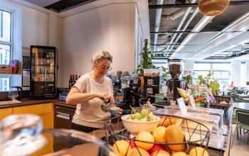 Binnenzicht van kantoorruimte op Fluwelen Burgwal 58, Den Haag Centrum, met een persoon die koffie zet bij een pantry, omringd door fruit en planten.