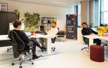 Zakelijke omgeving op Waldopstraat 5, Den Haag Centrum, met mensen in gesprek in een moderne kantoorruimte. De ruimte is voorzien van bureaus en enkele planten.