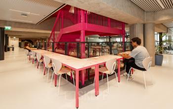 Moderne kantoorruimte in Den Haag Centrum met lange tafel, roze accenten en mensen die werken op laptops onder opvallende trappen. Geschikt voor kantoorruimte huren.