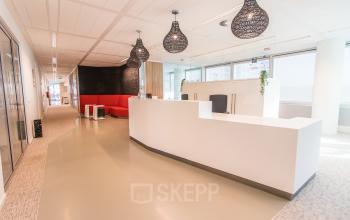Moderne kantoorpand lobby in Den Haag met strakke witte balie en rode zithoek. Ruime en lichte kantoorruimte huren met glaswanden. Ideaal voor bedrijven die een professionele uitstraling wensen.