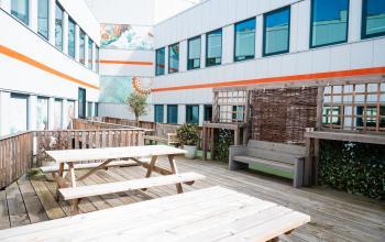 Een ruime buitenplek bij een kantoorpand op Tauber 52 in Den Haag Leidschenveen-ypenburg, met houten picknicktafels omgeven door moderne architectuur en kleurrijke muurschilderingen. Ideaal voor huur als ontspannen werkruimte.