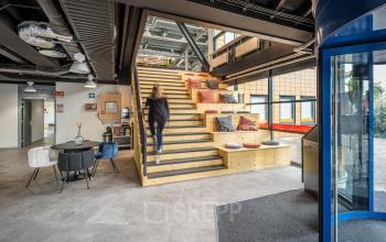 Kantoor gelegen op Tauber 52 in Den Haag Leidschenveen-Ypenburg met een moderne entree en zitruimte. Een persoon loopt de houten trap op, omringd door een informele werk- en overlegomgeving.