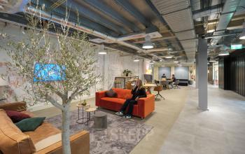 Binnenzicht van modern kantoorpand op Tauber 52, Den Haag Leidschenveen-Ypenburg, met kleurrijke meubels en mensen die druk aan het werk zijn. Ideaal voor bedrijven die kantoorruimte huren zoeken met een creatieve sfeer.