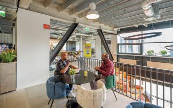 Moderne kantoorruimte huren in Den Haag Leidschenveen-Ypenburg, met industriële plafonds en kleurrijke stoelen. Twee mannen in gesprek aan een tafel, gelegen op Tauber 52.