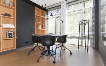 Kantoorruimte huren op President Kennedylaan 19 in Den Haag; compacte vergaderingstafel met vier stoelen, een staande lamp en ingebouwde planken bij het raam met jaloezieën.