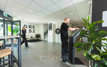 Kantoorruimte op Keulenstraat 9, Deventer met moderne ontvangsthal. Twee personen zijn bezig met zakelijke activiteiten: een staat bij een scherm en een ander loopt door de ruimte.