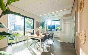 Kantoorruimte huren op Keulenstraat 9 in Deventer, met een lichte, moderne werkruimte gevuld met een grote tafel, stoelen en decoratieve elementen bij grote ramen. Geschikt voor zakelijke activiteiten en vergaderingen.