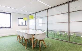 Moderne kantoorruimte huren aan de Zutphenseweg 29 in Deventer met grote ramen, groene vloerbedekking, glazen wanden en een lange tafel met stoelen perfect voor vergaderingen.