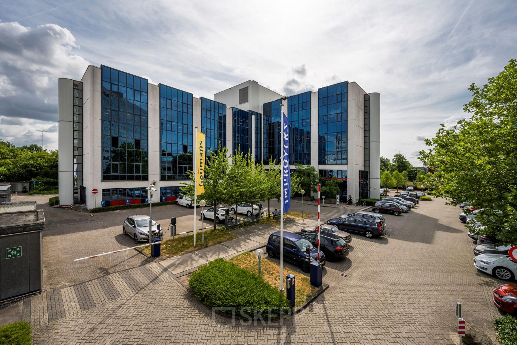 Het kantoorpand Diemermere 11-19 in Diemen met moderne glazen gevel en ruime parkeerplaats voor het gebouw.