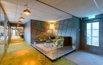 Moderne loungehoek in kantoorpand op Mijlweg 61, Dordrecht, met stijlvolle zitbanken en decoratieve muurkunst. Personen op de achtergrond bezig met zakelijke activiteiten in aangrenzende kantoorruimte.