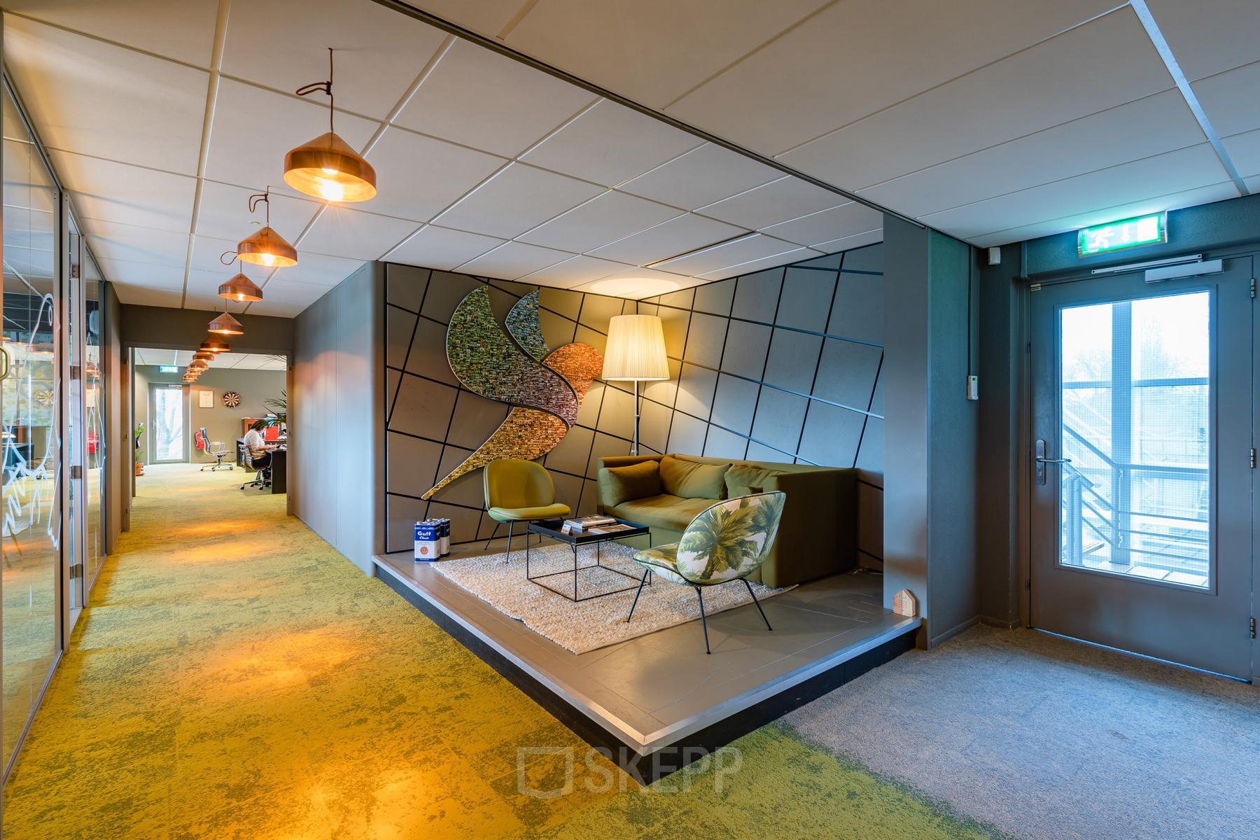 Moderne loungehoek in kantoorpand op Mijlweg 61, Dordrecht, met stijlvolle zitbanken en decoratieve muurkunst. Personen op de achtergrond bezig met zakelijke activiteiten in aangrenzende kantoorruimte.