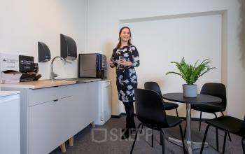 In een kantoorpand op Galvanistraat 7, Ede, ziet men een compacte pantry met een tafel en stoelen. Een vrouw is bezig koffie te zetten met een glimlach, wat zorgt voor een gastvrije sfeer.