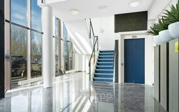 Entree van een modern kantoorpand op Celsiusstraat 34 in Ede, met een glazen pui, marmeren vloer en een glazen trap met blauwe bekleding. Kantoorruimte huren mogelijk.
