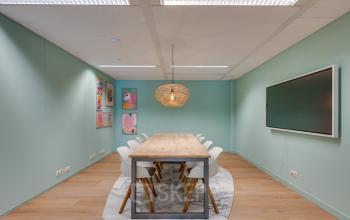 Vergaderruimte met lange houten tafel en moderne stoelen op Stadhuisplein 10 in Eindhoven Centrum. Kleurrijke kunstwerken aan de muur en een grote monitor maken deze plek geschikt voor kantoorruimte huren.