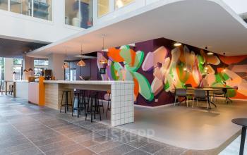Moderne kantoorruimte op Stadhuisplein 10, Eindhoven Centrum, met kleurrijke muurschilderingen. De ruimte is ingericht met tafels, stoelen en een bar, ideaal om te huren voor creatieve werksessies.