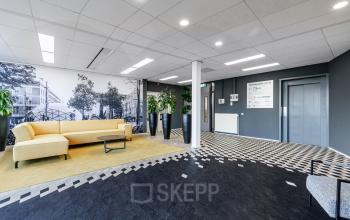 Een moderne kantoorlobby met een beige bank, planten en muurdecoraties in een gebouw op Meerenakkerplein 1-10, Eindhoven De Hurk. Ideaal voor kantoorruimte huren in dit kantoorpand.