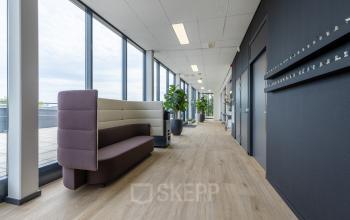Moderne kantoorruimte huren in Eindhoven De Hurk met een smalle gang, voorzien van glazen wanden, houten vloer en zitmeubilair. Veel lichtinval en planten sieren het strakke interieur.