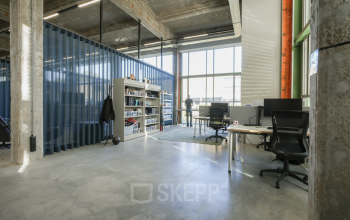 Kantoorruimte huren in modern kantoorpand op Frederiklaan 10A, Eindhoven Strijp-S. Industriële uitstraling met grote ramen, glazen wanden, werkplaatsen met bureaus en stoelen. Persoon aan het werk met natuurlijk lichtinval.