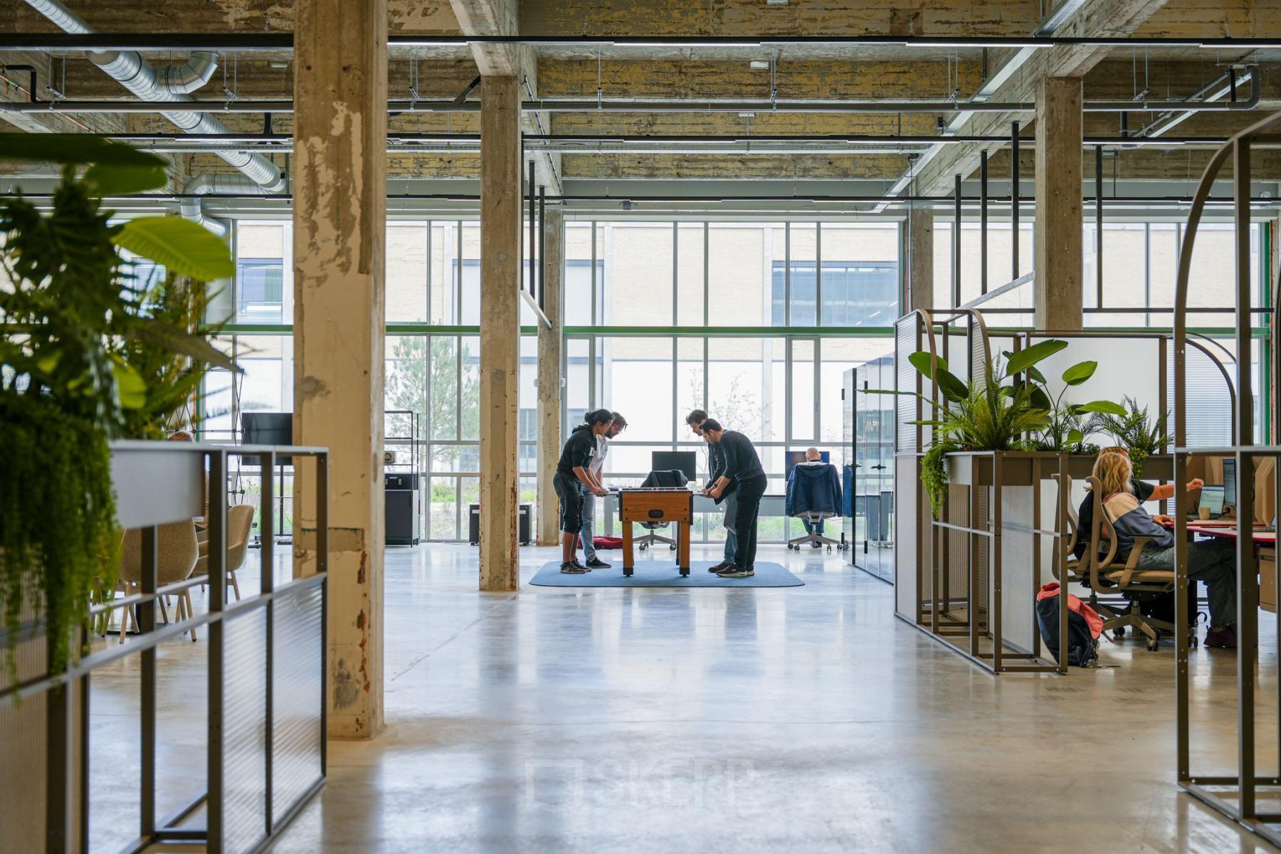 Industriële kantoorruimte huren aan de Frederiklaan 10A in Eindhoven Strijp-S met open vloerplan. Mensen werken en spelen tafelvoetbal in een modern, groen ingerichte setting.