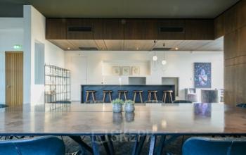 Moderne kantoorruimte met houten accenten en een lange tafel in Eindhoven Strijp-S. Achterin is een keuken zichtbaar met hoge krukken. Ruimte voor kantoorruimte huren.