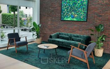 Ontspannen lounge met een groene bank, twee grijze stoelen en planten op Hoenderkamp 20, Emmen. Ideaal voor wie kantoorruimte wil huren. De lichte ruimte biedt een uitnodigende sfeer.