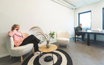 Twee mensen in een gemeubileerd kantoorpand op Brouwerijstraat 1, Enschede. Een persoon zit aan een bureau met een laptop, terwijl de ander in een fauteuil telefonerend is. Ideaal voor kantoorruimte huren.
