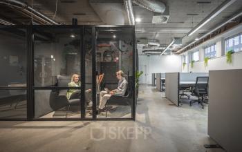 Moderne kantoorruimte huren op Brouwerijstraat 1 in Enschede. Medewerkers voeren een zakelijk overleg in een glazen vergaderruimte, terwijl andere werkplekken verder zijn ingericht voor productiviteit.