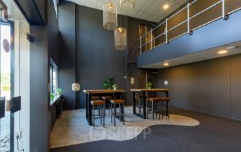 Kantoorruimte in Enschede bij Marssteden 20 met hoge plafonds, een moderne zithoek met tafels en krukken, en voldoende lichtinval. Ideaal voor bedrijven die een kantoorpand willen huren.