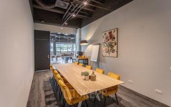 Vergaderruimte in kantoorpand op Wethouder Beversstraat 185, Enschede, met lange tafel en gele stoelen, kunstwerk aan muur en raam naar open werkruimte.