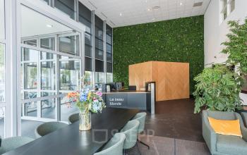 Moderne kantoorpandlobby op Brandweerstraat 20-30 in Enschede met groene muur, receptiebalie, comfortabele stoelen, en een lange tafel met bloemen. Perfecte ruimte voor dynamische zakelijke ontmoetingen.