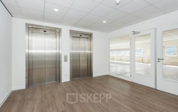 Moderne entree van kantoorpand op Brandweerstraat 20-30, Enschede met stijlvolle houten vloer en strakke, witte muren. Twee liften en glazen deur geven toegang tot verdere kantoorruimte.