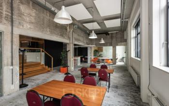 Kantoorruimte huren in Brouwerijstraat 10, Enschede met houten tafels, rode stoelen, grijze industriële muren, en grote ramen. Achterin is een glazen vergaderzaal zichtbaar.