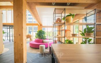 Moderne kantoorruimte op Moutlaan 1-23, Enschede met houten structuren, kleurrijke banken en groene planten. Ideaal voor wie een inspirerende kantoorpand wil huren met een uitnodigende lounge sfeer.