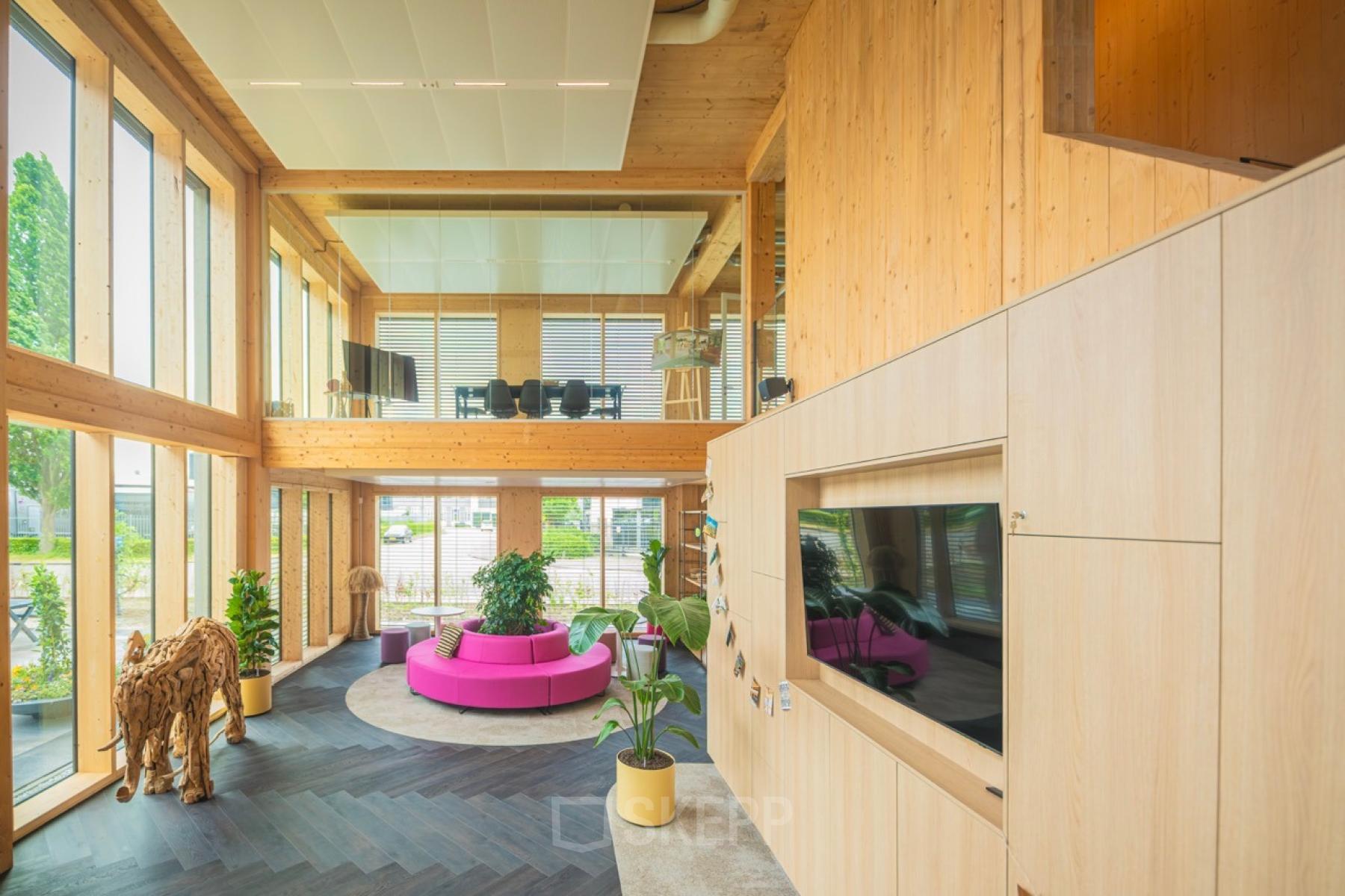 Lichte en moderne kantoorruimte met houten accenten en grote ramen op Moutlaan 1-23, Enschede. Roze bank en planten verlevendigen het interieur. Ideaal voor werk en vergaderingen.