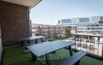 Uitzicht vanaf een balkon van kantoorpand Kuipersdijk 2-4, Enschede, met houten picknicktafels en uitzicht op omliggende gebouwen en parkeerplaats. Ideaal voor een informele pauze tijdens het werk.